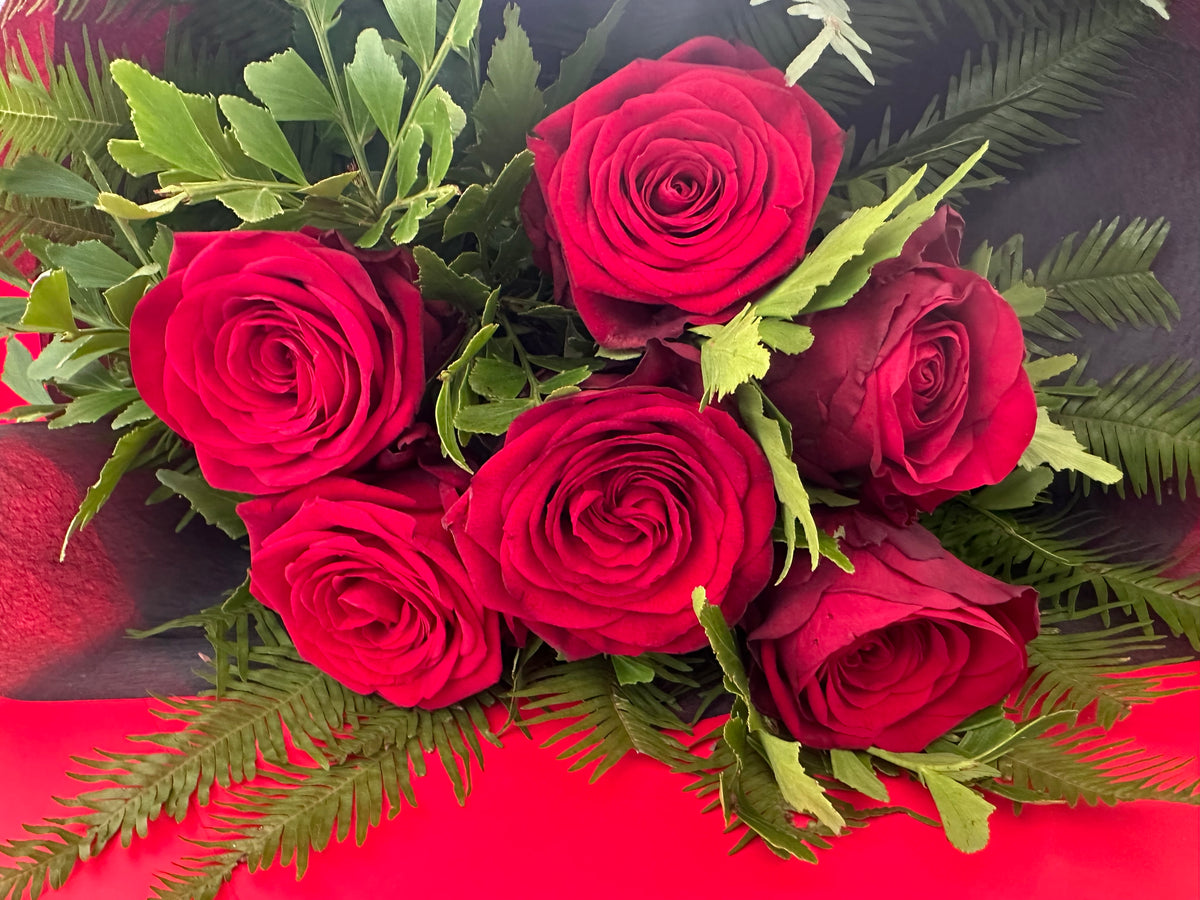 A bouquet of six deep red roses with green leaves and fern-like foliage, with red and black wrap