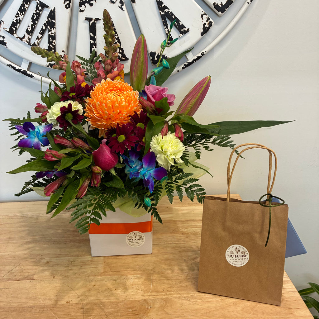 Floral arrangement of various brightly coloured blooms and gift bag.