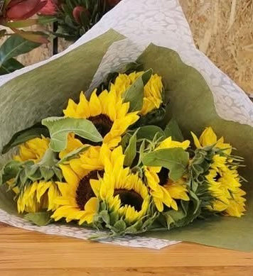 A bouquet of bright yellow sunflowers with green leaves, wrapped in white patterned paper and green tissue, resting on a wooden surface. Delivered to Sweet Brew Lilydale 