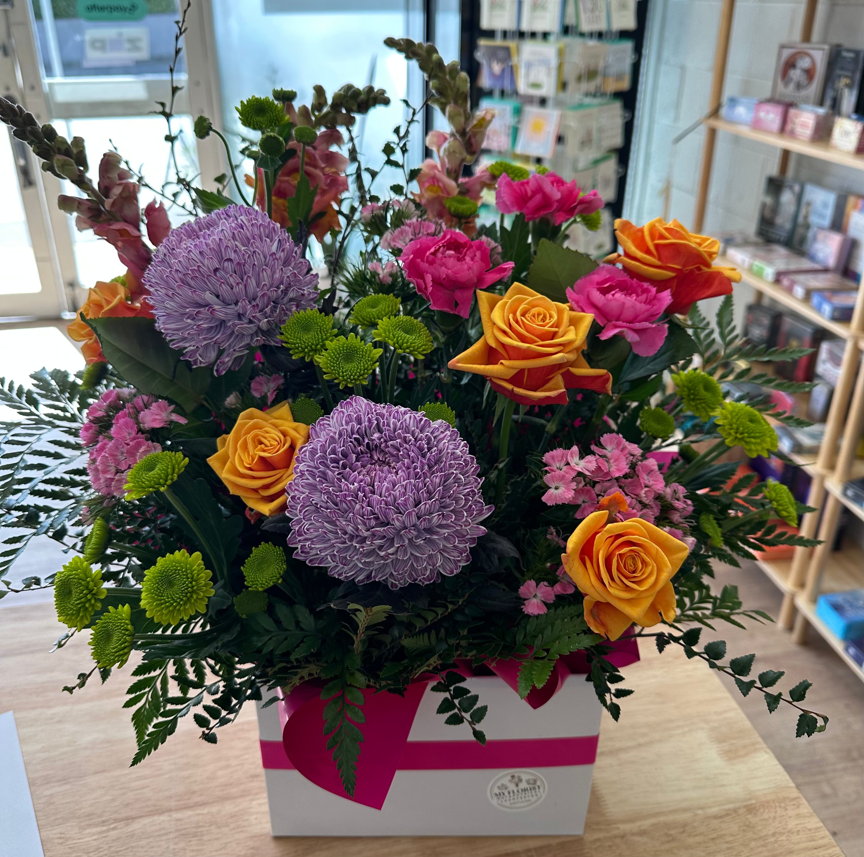 A vibrant mixed floral arrangement in a white gift box with a pink ribbon, displayed on a wooden table in a shop setting. The bouquet features prominent purple chrysanthemums, pink roses, orange roses, and green foliage.