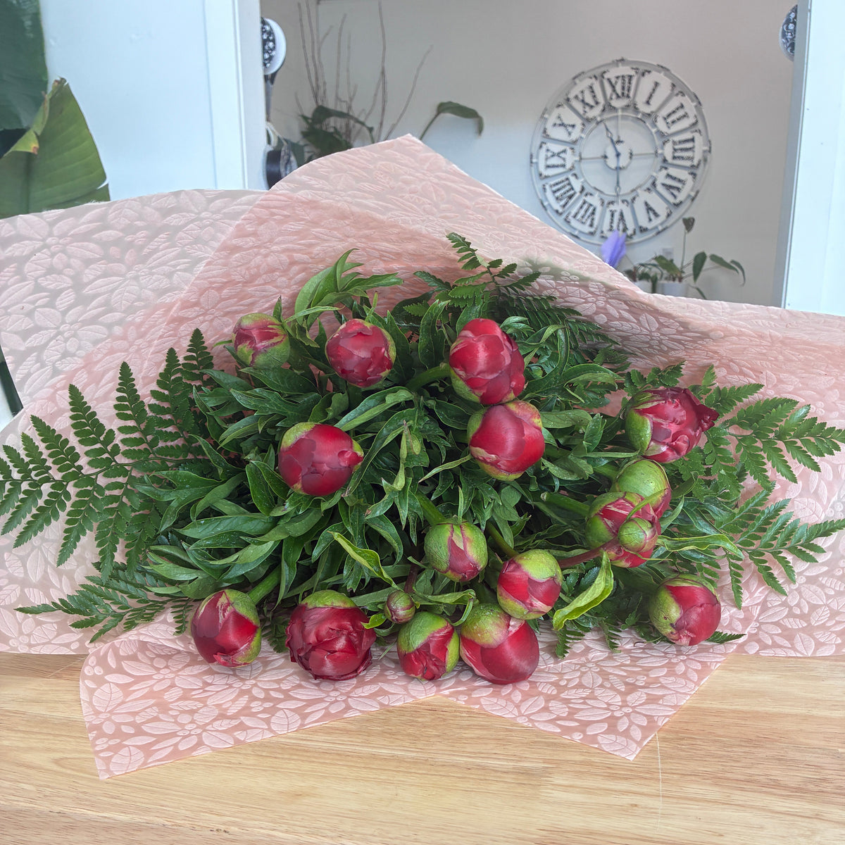 A bouquet of red peony flower buds with green leaves and fern accents, wrapped in pink patterned paper and placed on a wooden surface. Launceston florist delivery