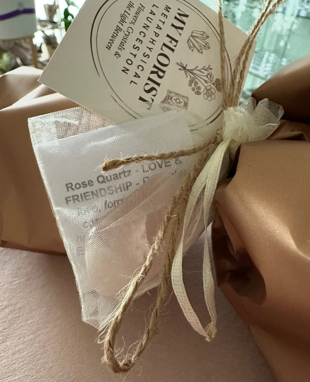 A close-up of a wrapped flower gift  with brown paper wrapping, a cream ribbon tied in a bow, a small sheer white pouch printed with text, and a round card reading 'MY FLORIST METAPHYSICAL LAUNCESTON'.